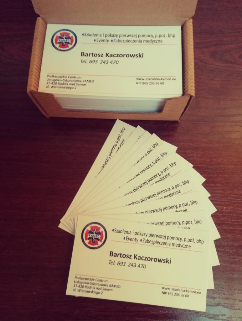 Wizytówki firmy oferującej szkolenia z pierwszej pomocy, ppoż, bhp oraz zabezpieczenia medyczne, ułożone w pudełku i na stole.