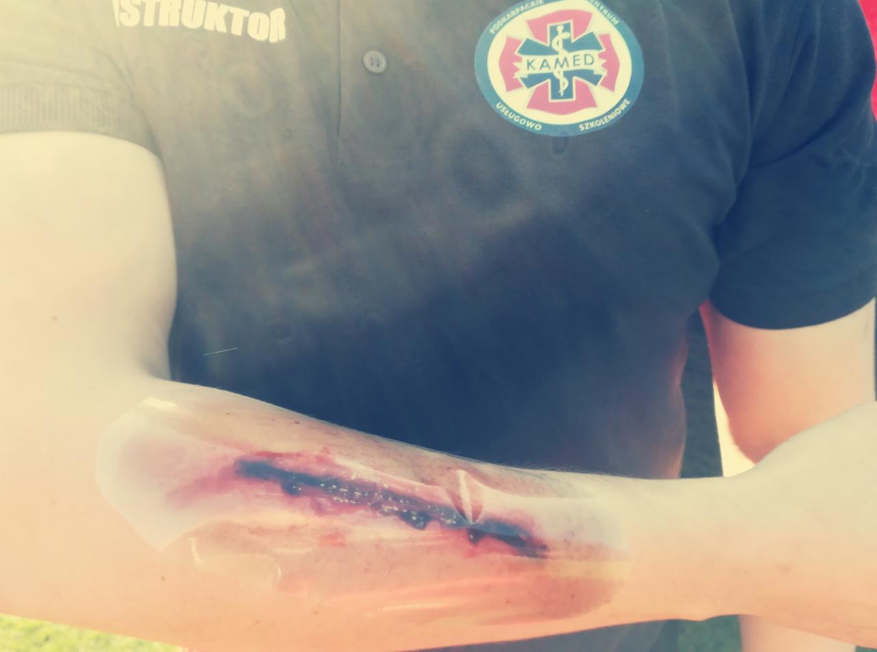 Ramię z realistyczną symulacją rany ciętej zabezpieczonej folią, widoczny instruktor w koszulce z logo centrum szkoleniowego.