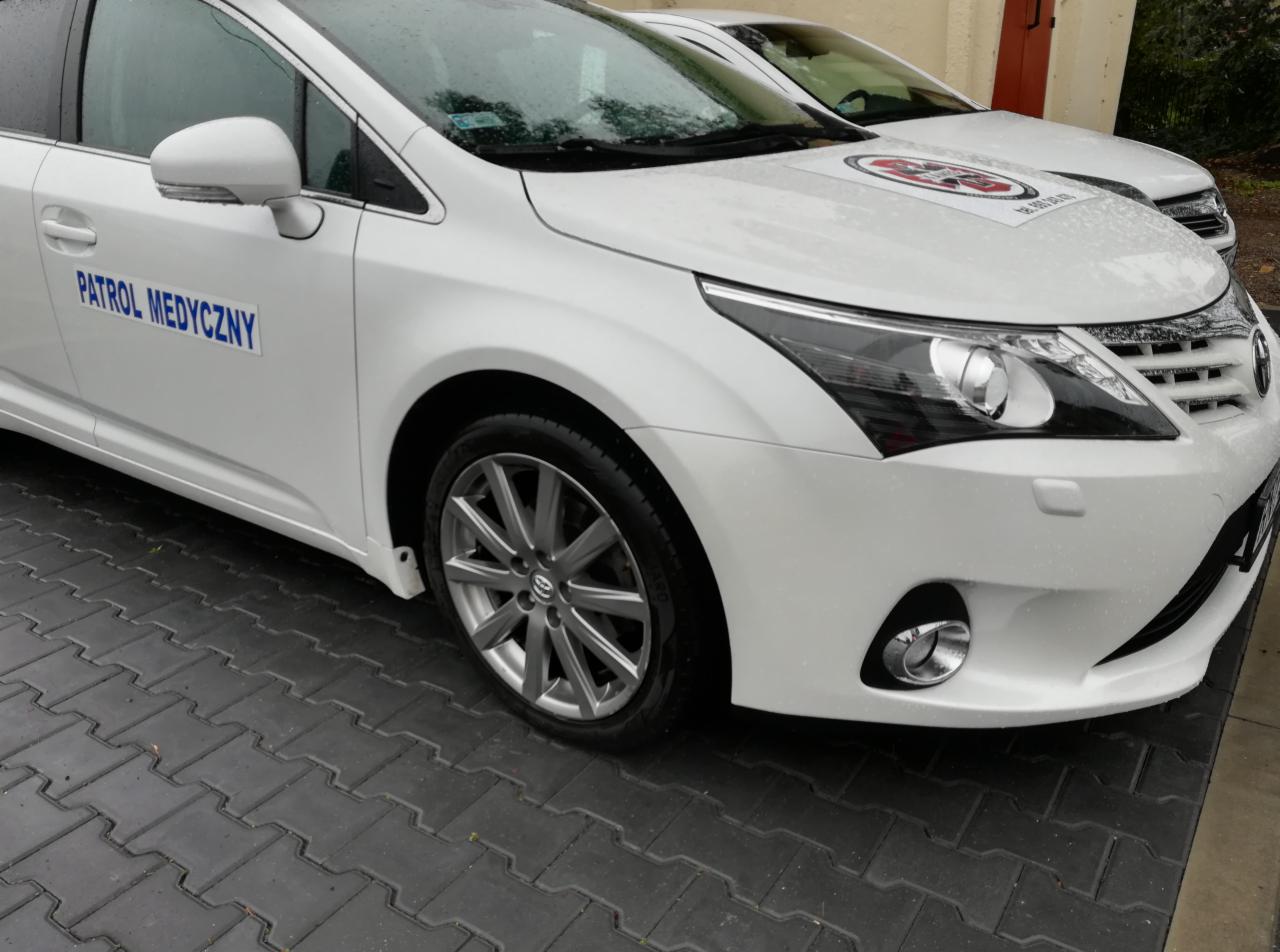 Biały samochód oznaczony jako 'Patrol Medyczny' zaparkowany na szarej kostce brukowej, widoczne krople deszczu na karoserii.