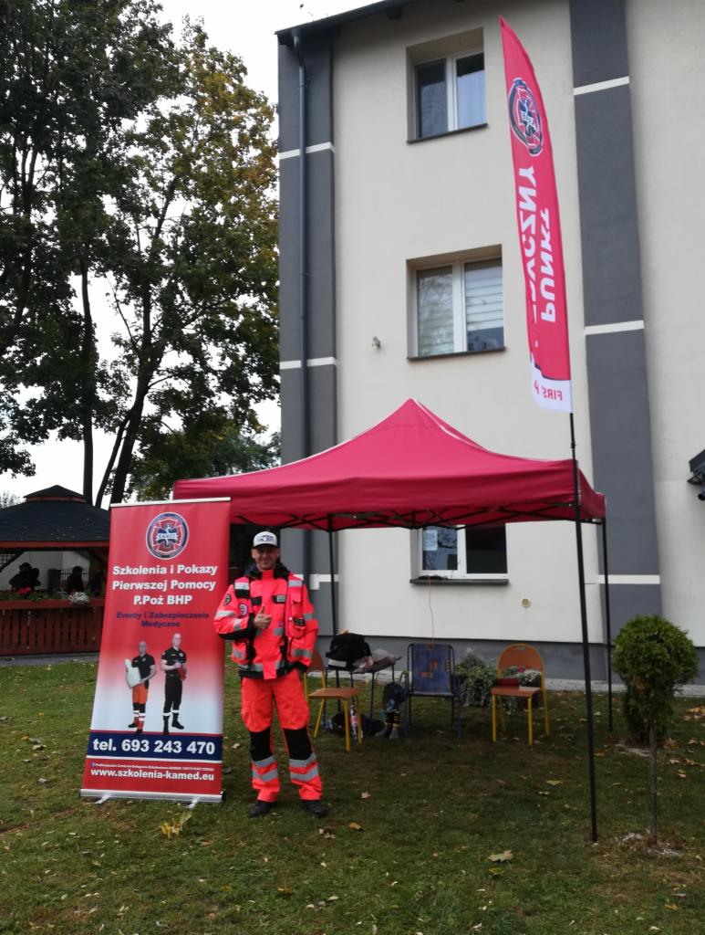 Instruktor w odblaskowym kombinezonie stoi obok baneru z informacją o szkoleniach z pierwszej pomocy i P.Poż BHP, pod czerwonym namiotem na trawniku przed budynkiem.
