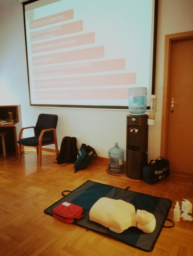 Sala szkoleniowa z manekinem do resuscytacji na macie, czerwona torba AED z widocznym napisem AED, butelki z płynem dezynfekcyjnym, krzesło, dystrybutor wody oraz rzutnik z wyświetlanymi punktami...