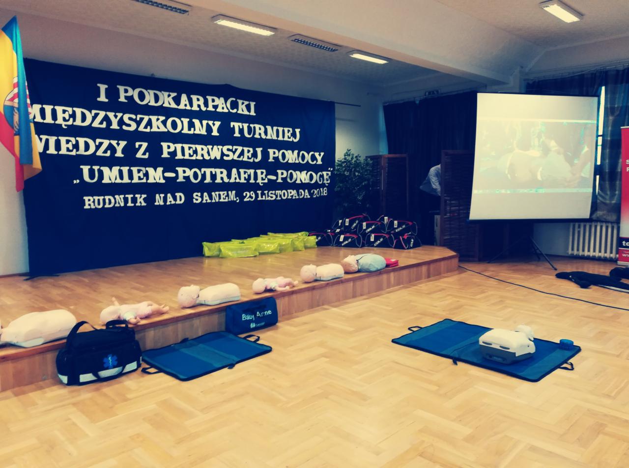 Sala z przygotowanym sprzętem do szkolenia z pierwszej pomocy: manekiny, torby ratunkowe, defibrylator na macie, baner informacyjny o turnieju wiedzy z pierwszej pomocy w tle oraz ekran projekcyjny.
