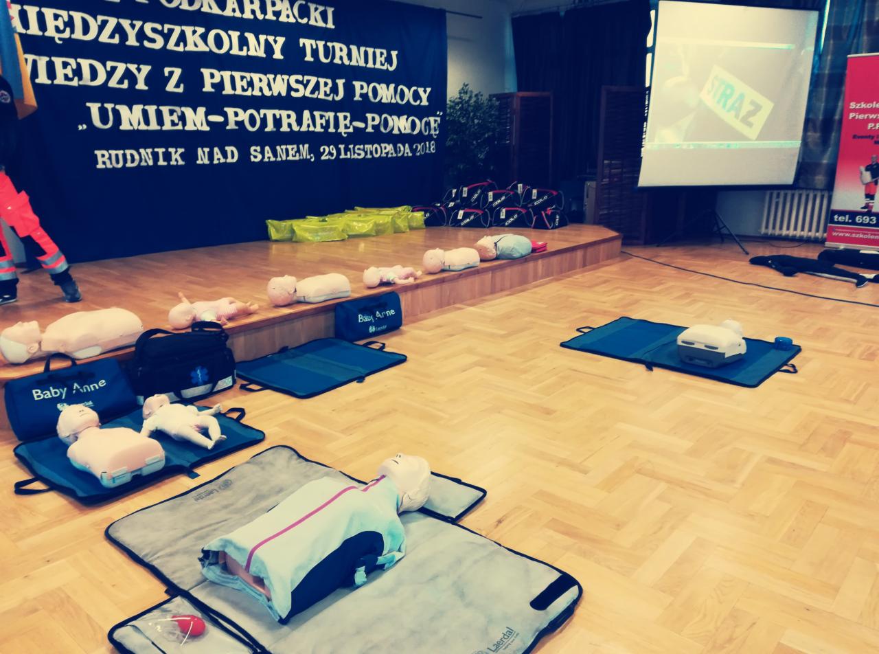 Sala z manekinami do nauki resuscytacji na podkładkach, sprzęt treningowy, plakat turnieju wiedzy o pierwszej pomocy w Rudniku nad Sanem w tle.