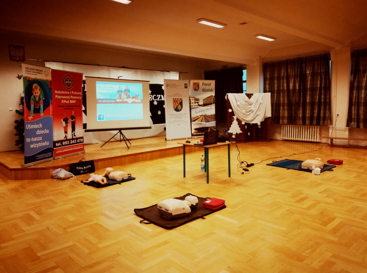 Sala szkoleniowa z manekinami do ćwiczeń resuscytacji niemowląt i dorosłych, rzutnik wyświetla prezentację, widoczne banery reklamowe organizatorów.