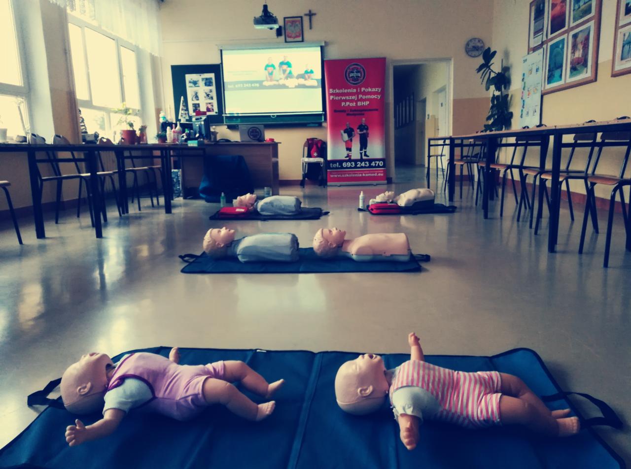 Sala szkoleniowa z manekinami do nauki resuscytacji, w tle rzutnik z prezentacją i baner reklamowy firmy szkoleniowej.