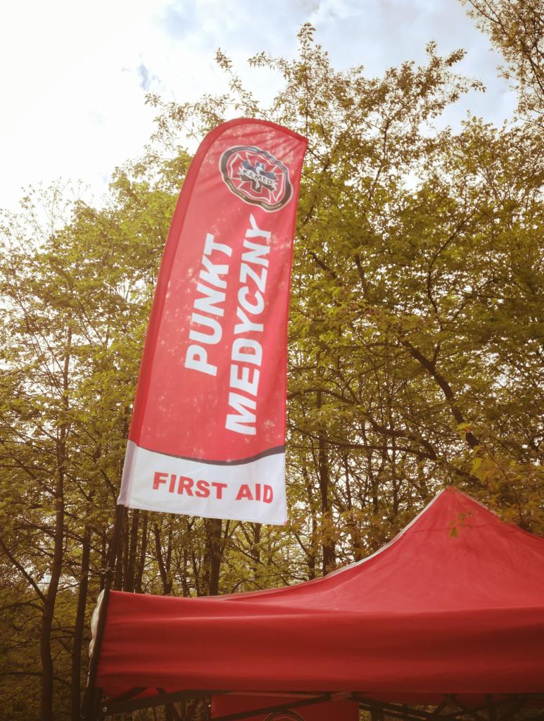 Czerwony, pionowy baner z napisem 'Punkt Medyczny' i 'First Aid' oraz logo służb ratowniczych, umieszczony przed czerwonym namiotem na tle drzew.