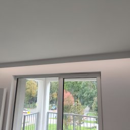 PremiumSolutions - wykończenia wnętrz mieszkań pod klucz - Minimalistyczny pokój z maskownicą karnisza i oświetleniem LED, widok na balkon i zieleń za oknem. Nowoczesne wykończenie wnętrz.