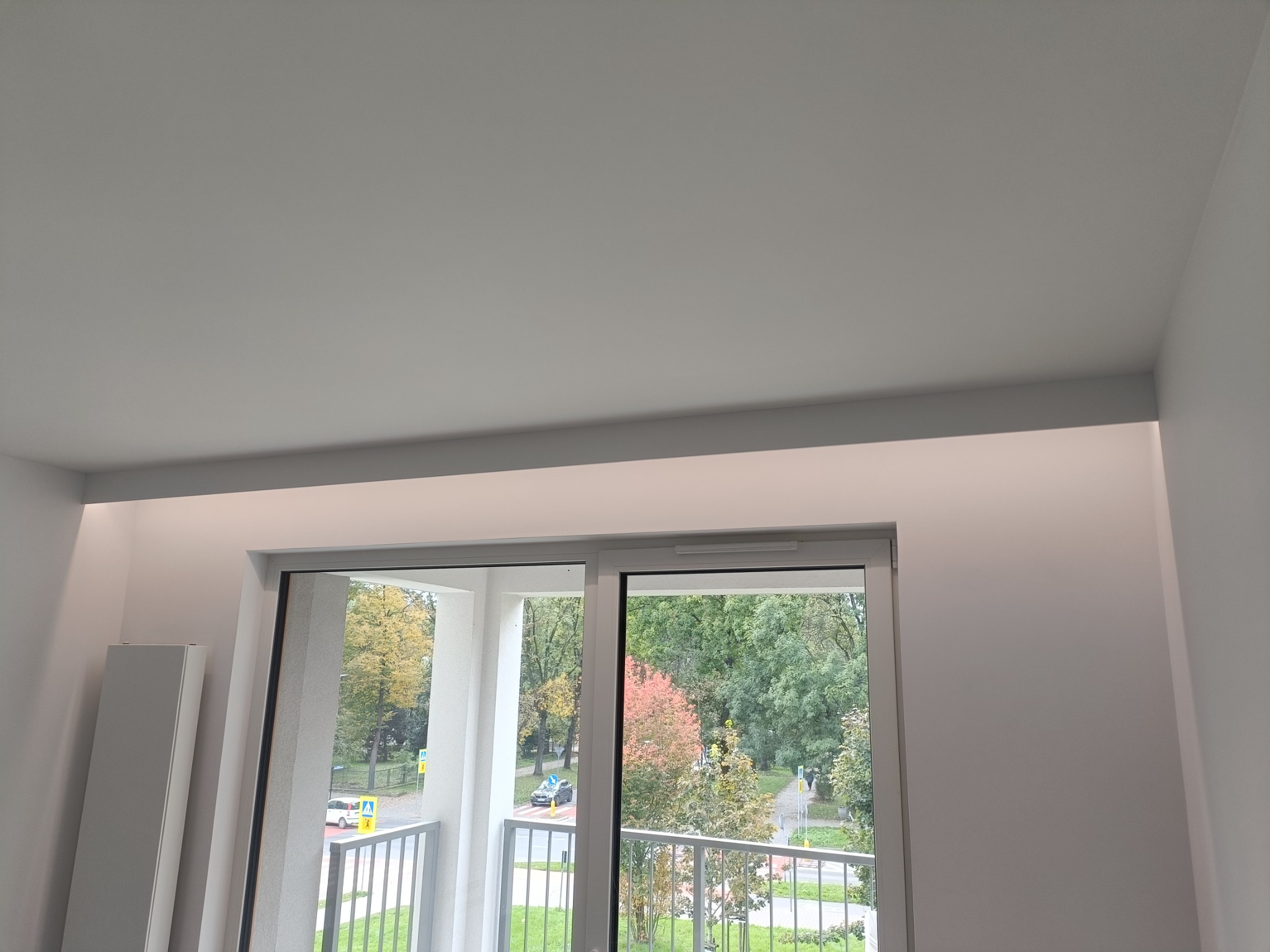 Minimalistyczny pokój z maskownicą karnisza i oświetleniem LED, widok na balkon i zieleń za oknem. Nowoczesne wykończenie wnętrz.