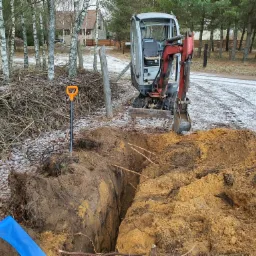 Mini koparka przy wykopie w ziemi, widoczne kable i rura, łopata wbita obok wykopu, w tle drzewa i częściowo ośnieżony teren.