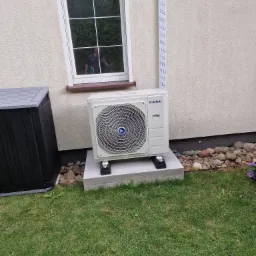 Zewnętrzna jednostka pompy ciepła Kaisai zamontowana na betonowym fundamencie obok okna z odbiciem fotografa, widoczna instalacja wzdłuż ściany budynku i fragment ogrodu.