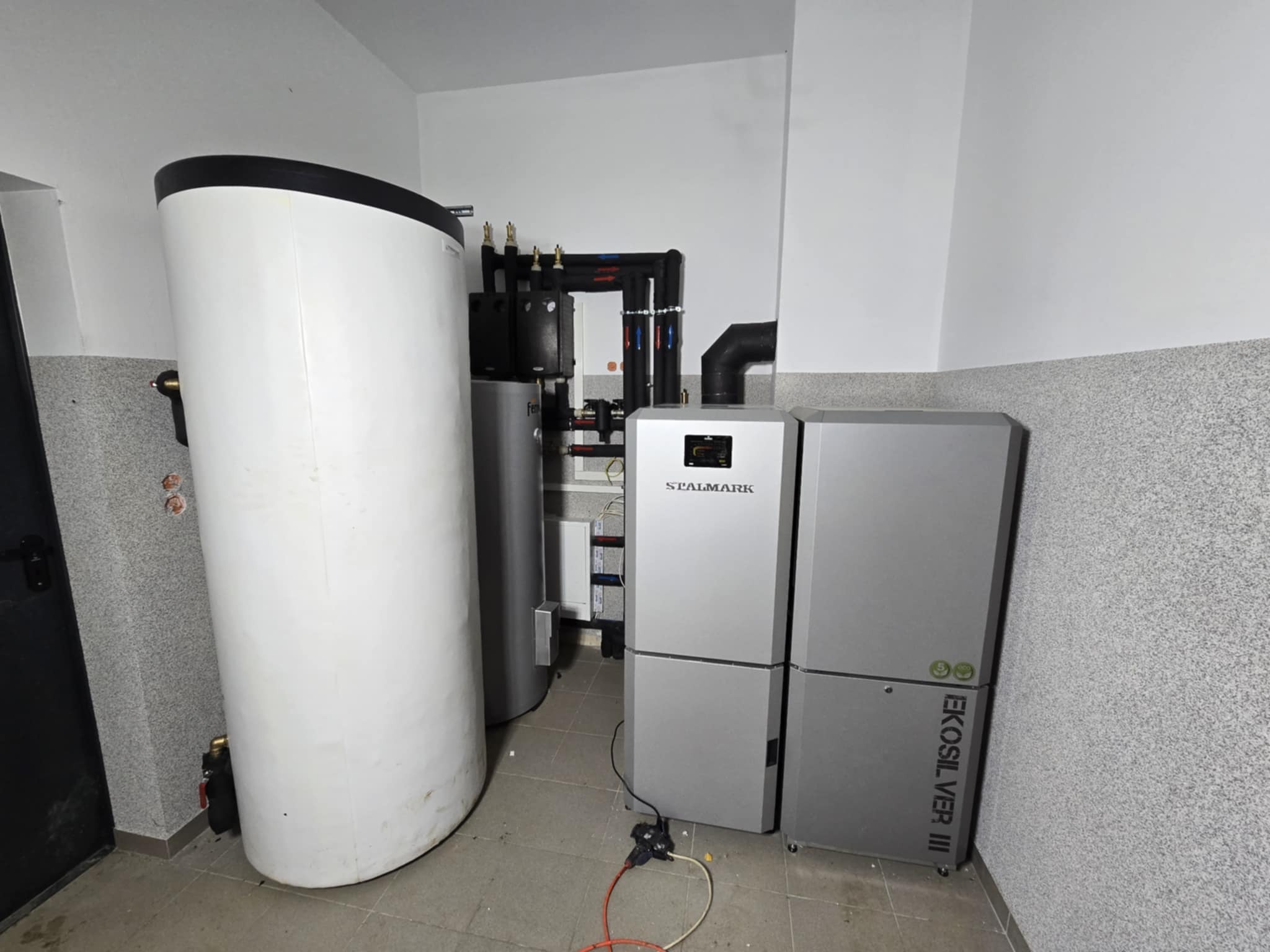 Kotłownia: biały zasobnik ciepłej wody z czarną górą, obok szary piec z napisem SEALMARK i model EKOSILVER III, widoczne rury i armatura, szara ściana w tle.