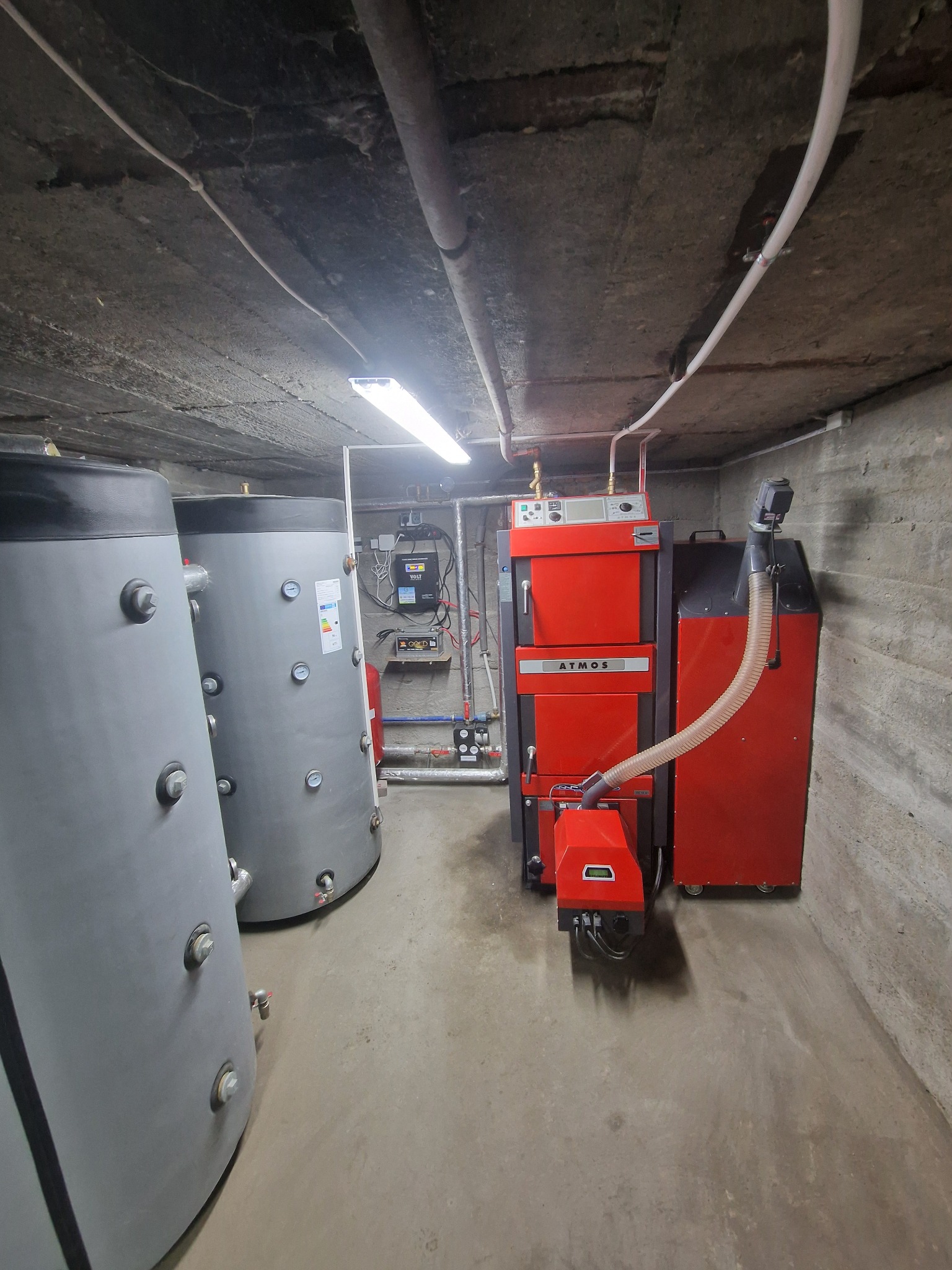 Kotłownia z dwoma szarymi zbiornikami akumulacyjnymi i czerwonym kotłem Atmos z podajnikiem, widok na instalację w piwnicy o betonowych ścianach i suficie.