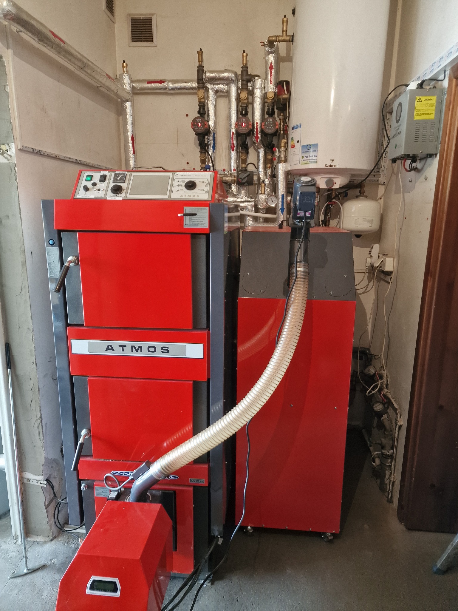 Czerwono-szary piec Atmos z podajnikiem, widoczne rury instalacji grzewczej, manometry i zbiornik wyrównawczy w kotłowni.