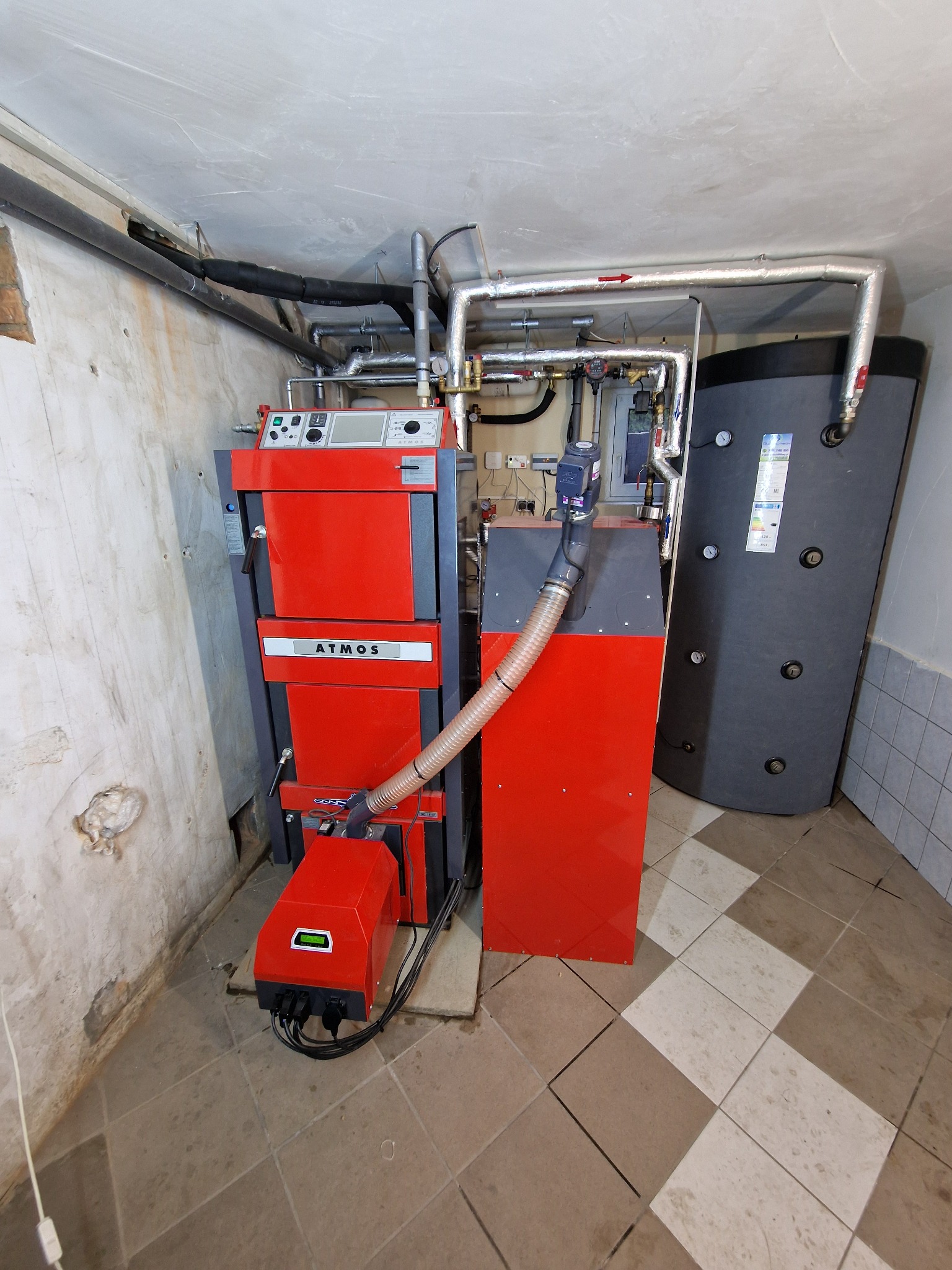 Czerwony piec ATMOS z podajnikiem i zbiornikiem buforowym w kotłowni z szaro-beżową podłogą i rurami grzewczymi pod sufitem.