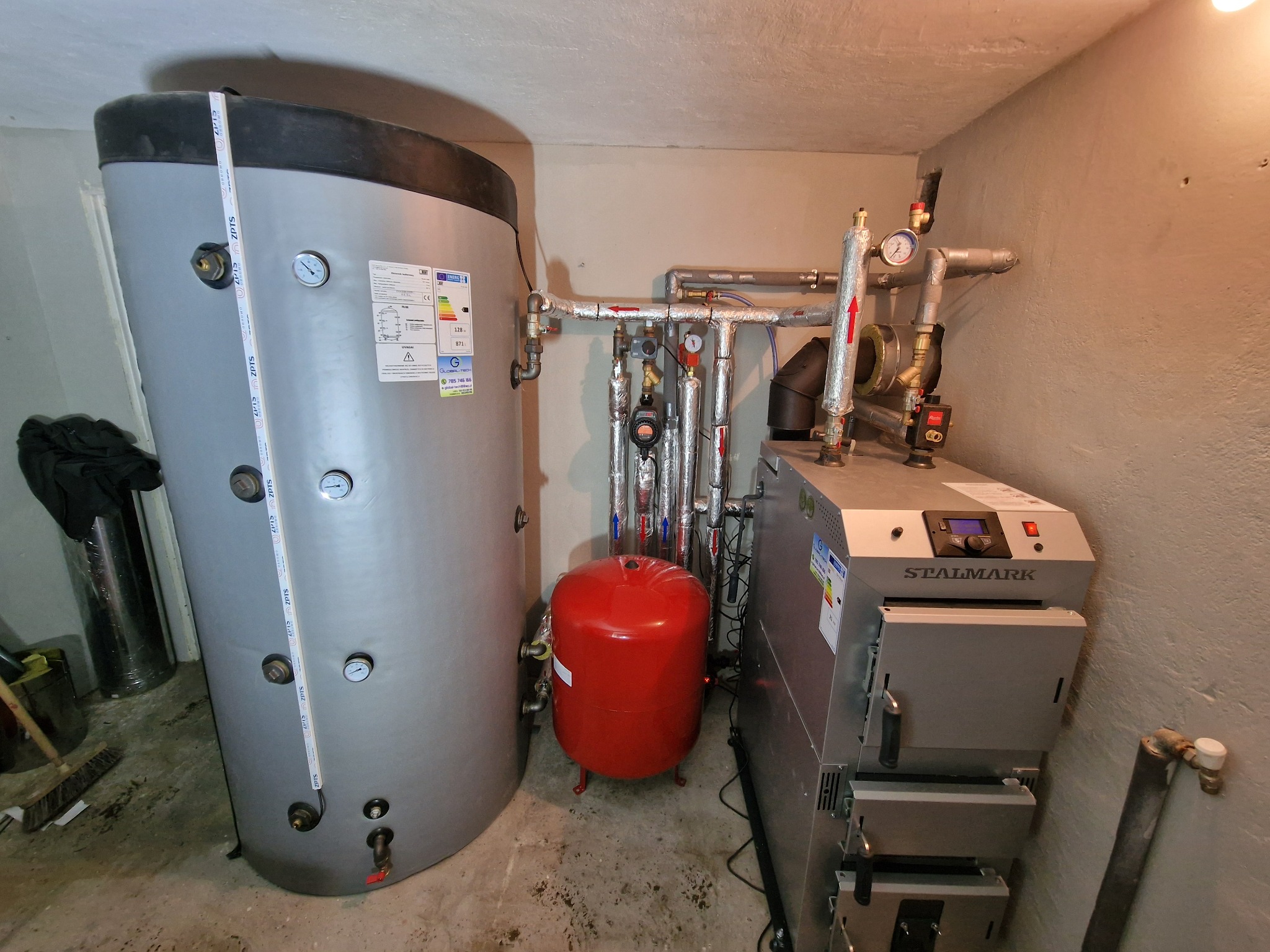 Szary zbiornik akumulacyjny ciepła z manometrami, czerwony zbiornik wyrównawczy i piec na paliwo stałe marki Stalmark w kotłowni.