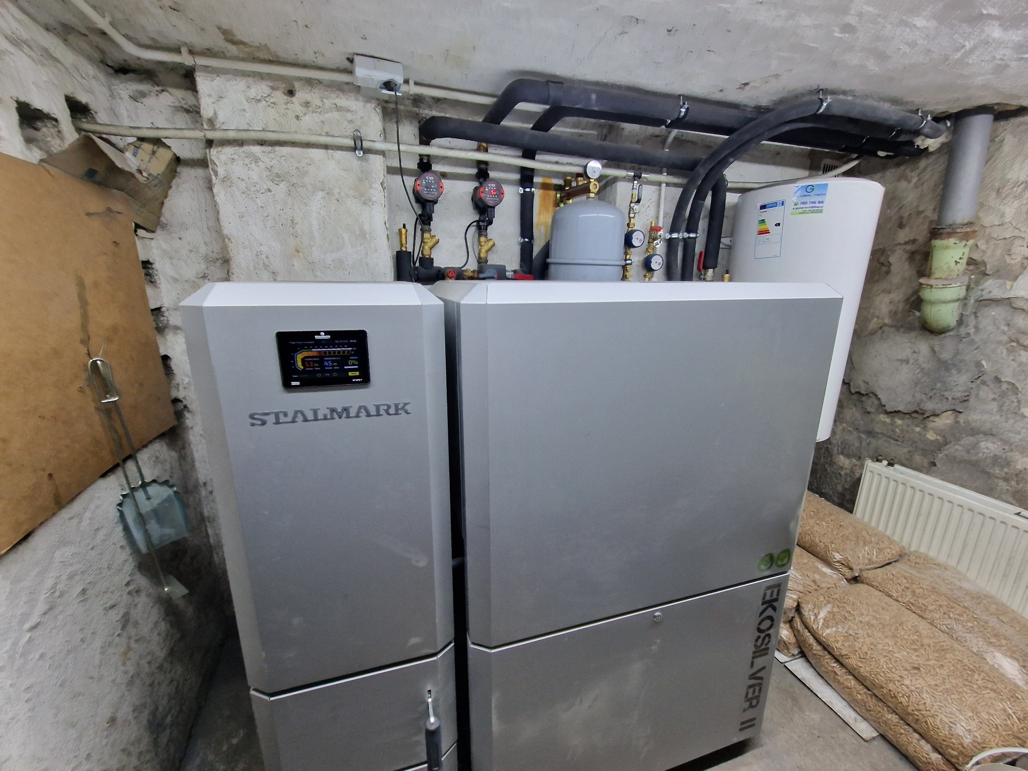 Nowoczesny piec CO Stalmark EkoSilver II w kotłowni, z widocznymi rurami instalacji grzewczej i zapasem pelletu obok grzejnika.
