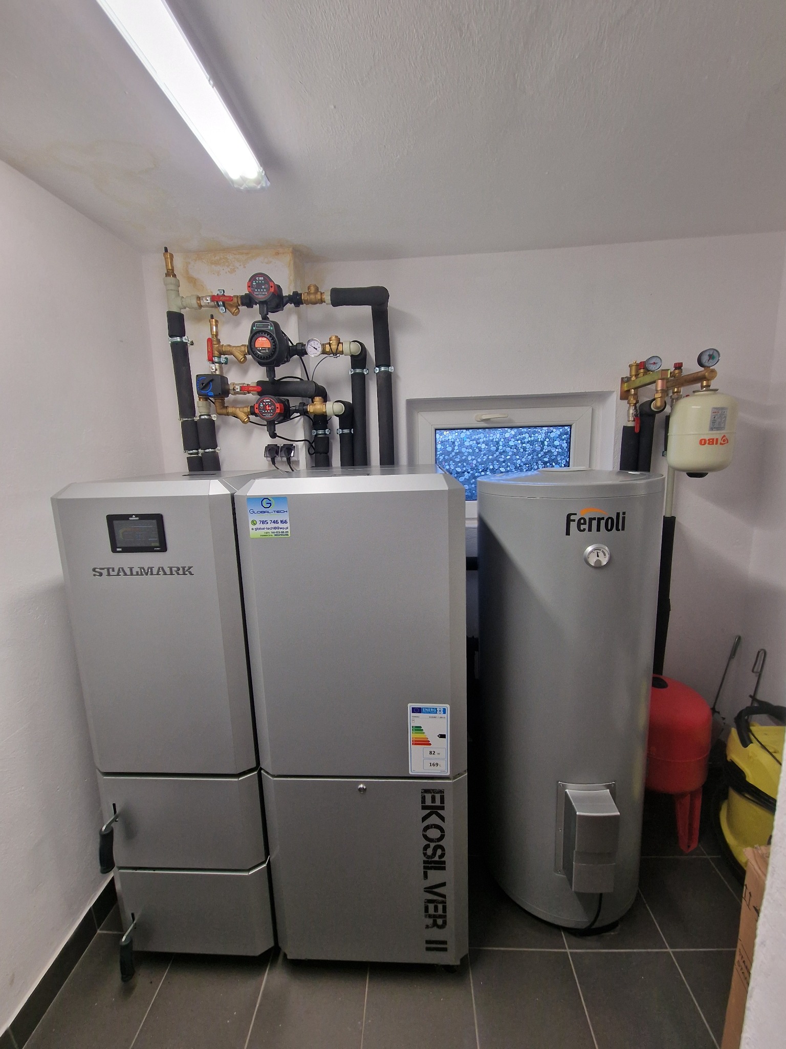 Zestaw urządzeń grzewczych w kotłowni: piec Stalmark z panelem sterowania, zasobnik Ferroli z osprzętem, instalacja hydrauliczna z pompami i zaworami na ścianie, widoczne rury i okno z ozdobną folią.