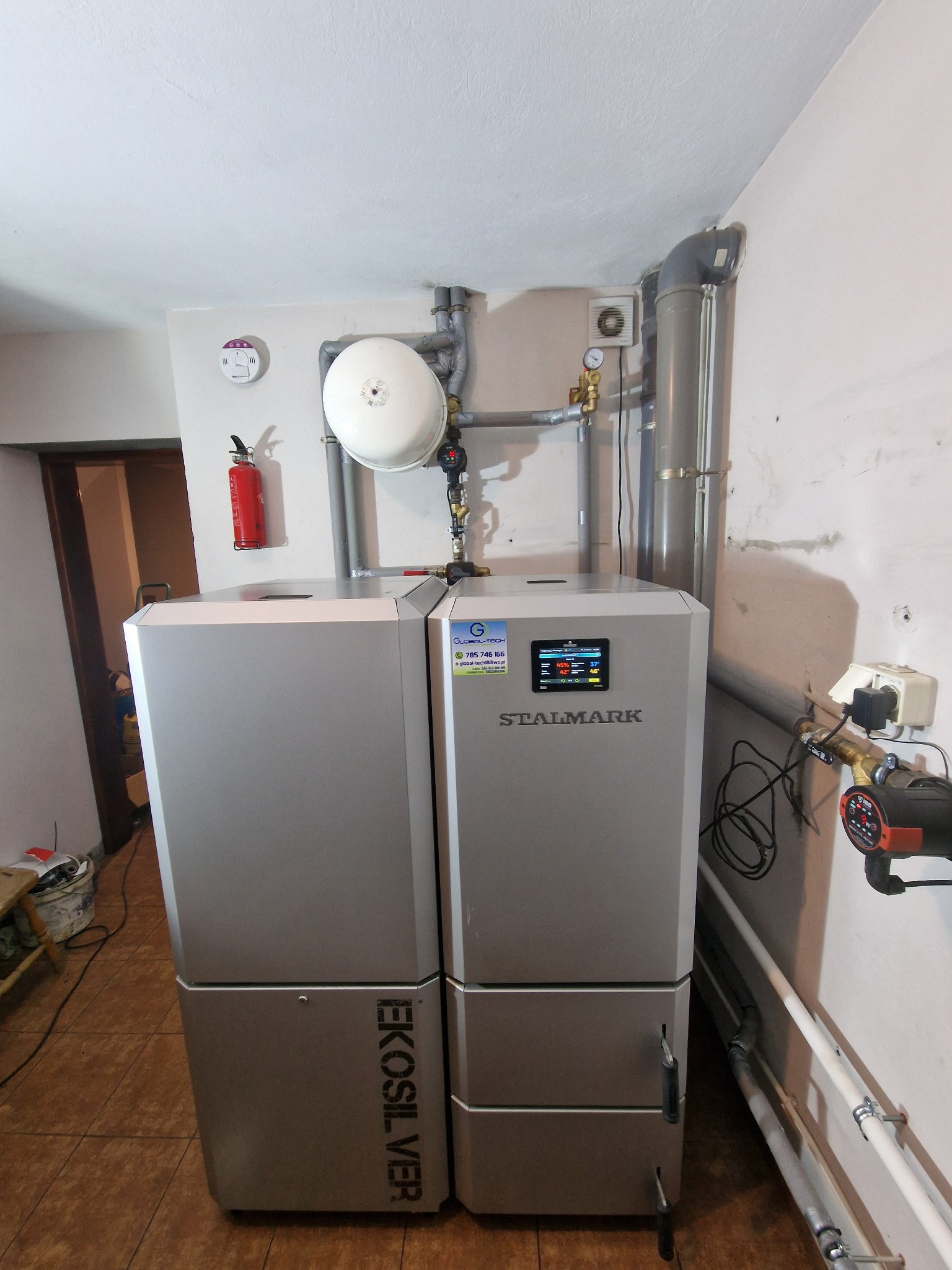 Dwa nowoczesne piece grzewcze Stalmark Ekosilver w kotłowni, widoczne rury instalacji, manometr i gaśnica na ścianie.