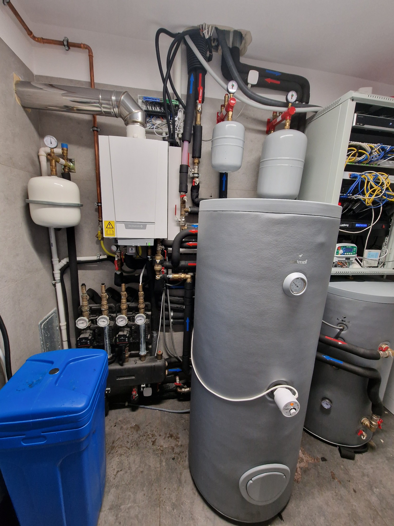 Kotłownia z piecem gazowym De Dietrich, dwoma szarymi zasobnikami ciepłej wody Galmet, rozdzielaczem hydraulicznym i niebieskim zmiękczaczem wody, widoczne liczne rury miedziane i izolowane.