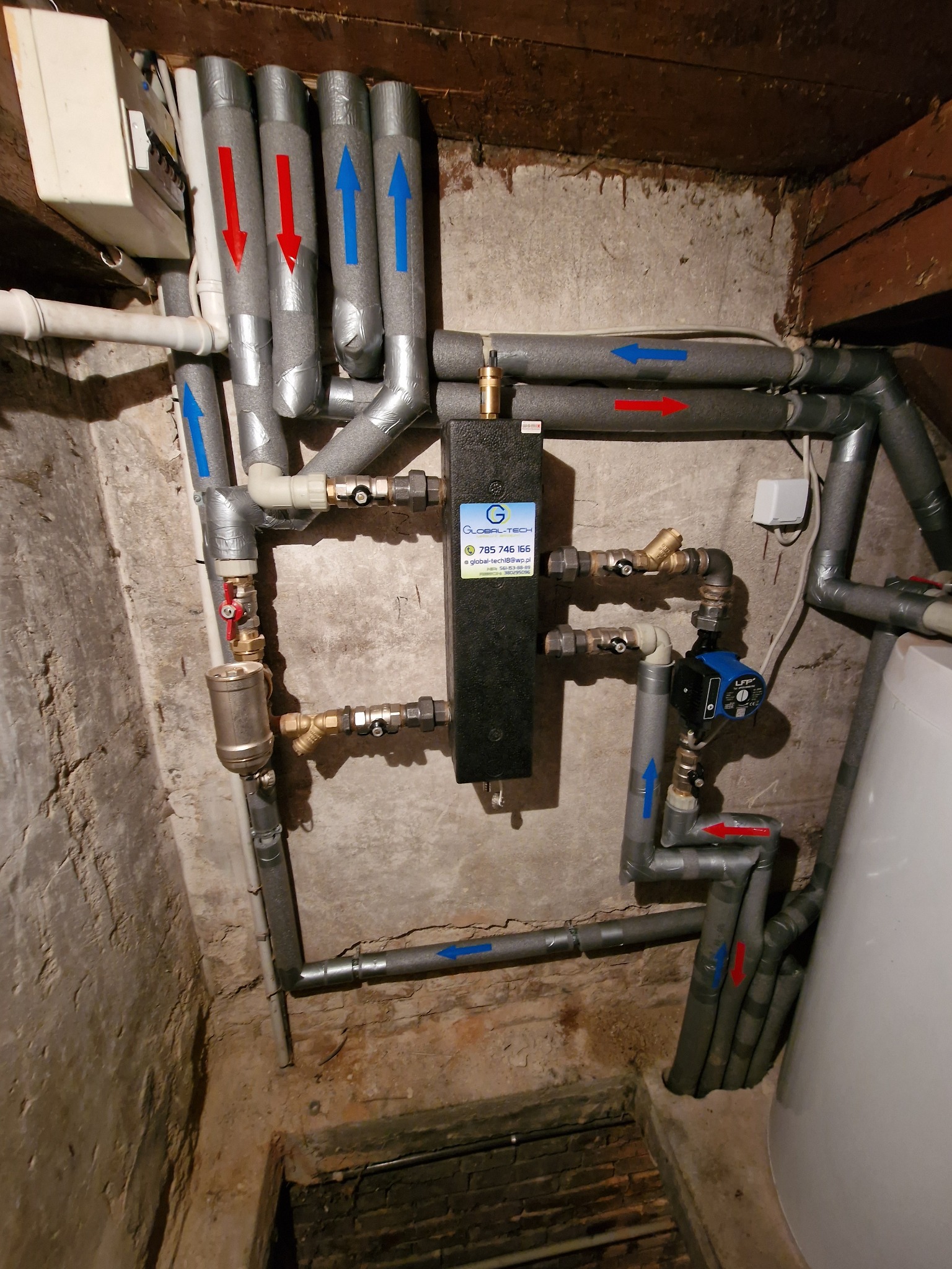 Złożony system rur grzewczych i hydraulicznych z izolacją, strzałkami wskazującymi przepływ, pompą obiegową LFP i filtrem, zamontowany na surowej ścianie w piwnicy z drewnianym stropem.