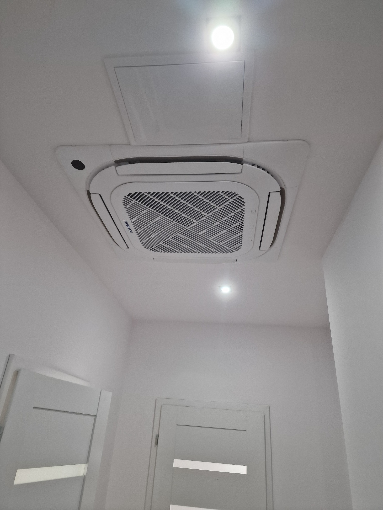 Kasetonowa jednostka klimatyzacyjna marki Kaisai zamontowana w suficie podwieszanym, widok od dołu z fragmentami dwóch białych drzwi w tle.