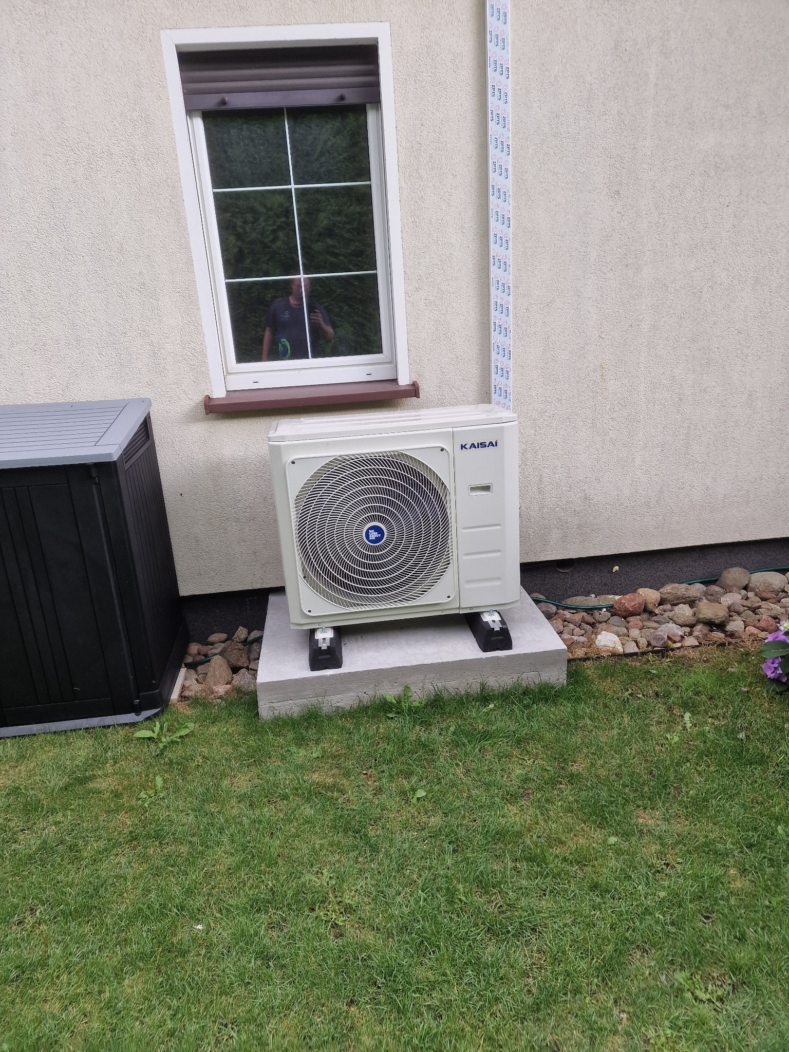 Zewnętrzna jednostka pompy ciepła Kaisai zamontowana na betonowym fundamencie obok okna z odbiciem fotografa, widoczna instalacja wzdłuż ściany budynku i fragment ogrodu.