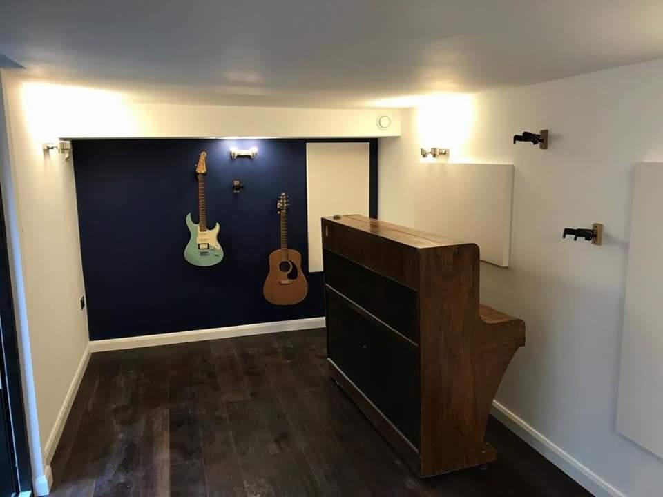 Wykończone pomieszczenie z ciemną podłogą, niebieską ścianą z dwiema gitarami i jasnymi panelami akustycznymi na białych ścianach, oświetlone lampami.