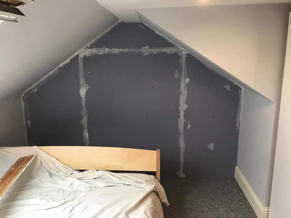 Montaż szarej okładziny wnęki na poddaszu, widoczne łączenia zabezpieczone taśmą, przykryte łóżko w pokoju.