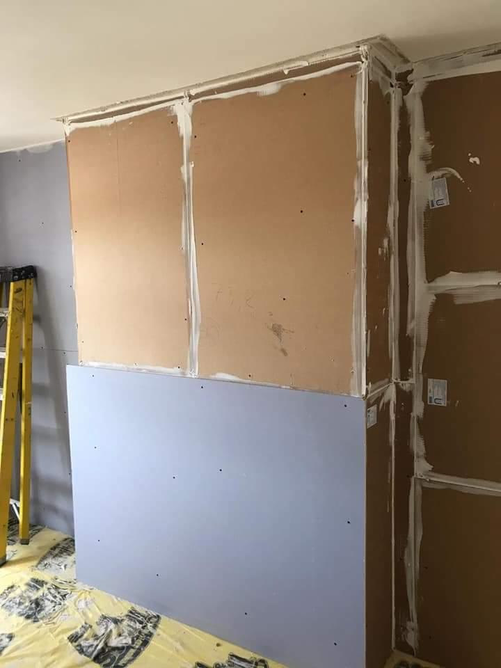 Montaż konstrukcji z płyt na ścianie, widoczne łączenia wypełnione masą szpachlową, żółta drabina oparta o ścianę, podłoga zabezpieczona folią ochronną.