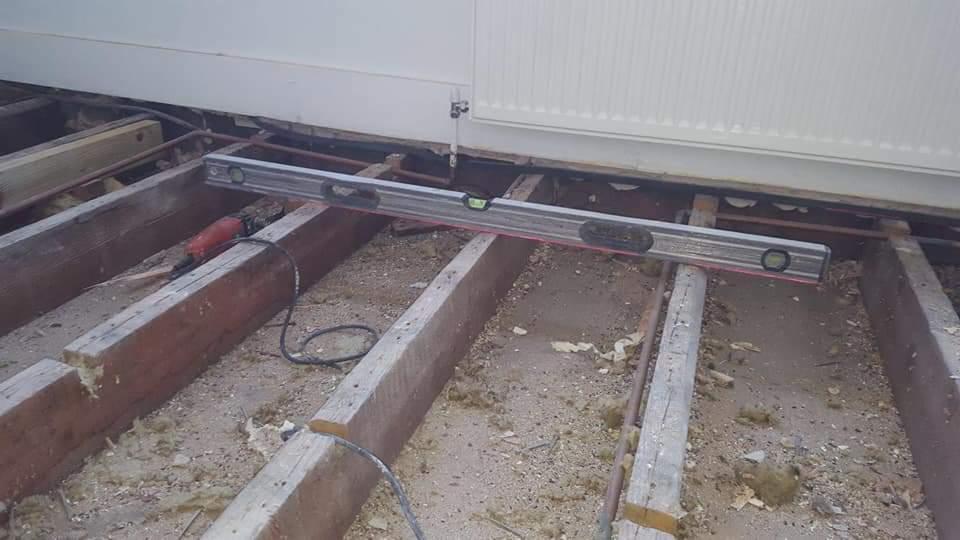 Poziomnica spoczywająca na drewnianej konstrukcji podłogi w trakcie remontu, widoczne rury i kabel od szlifierki.