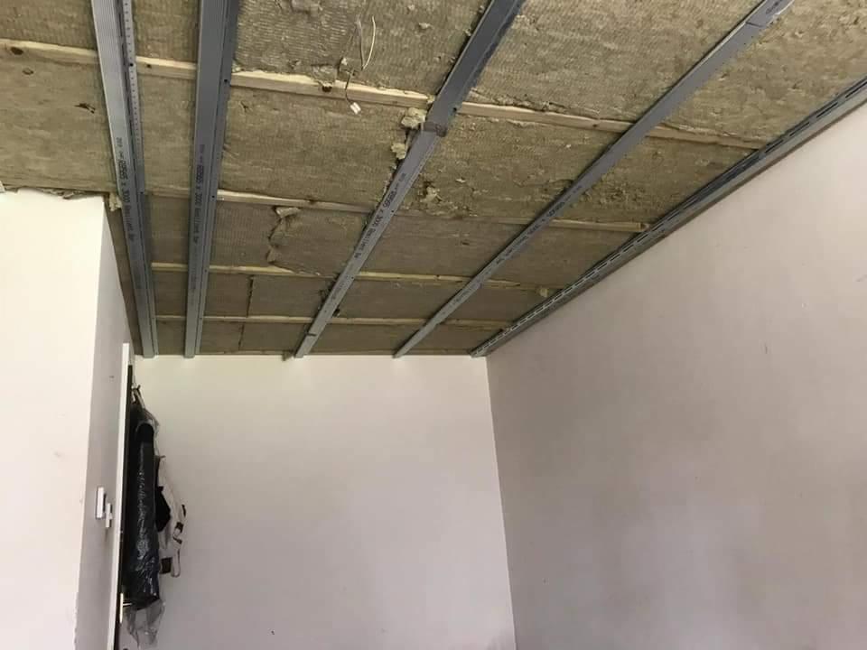 Montaż metalowego rusztu sufitowego z izolacją z wełny mineralnej, widoczne profile mocowane do stropu, przygotowanie do dalszych prac wykończeniowych.