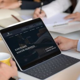 Studio pani B. Barbara Pisz - Tablet z wyświetloną stroną internetową kancelarii radców prawnych, widoczny fragment klawiatury i dłoń trzymająca kubek. W tle osoby przy biurku.