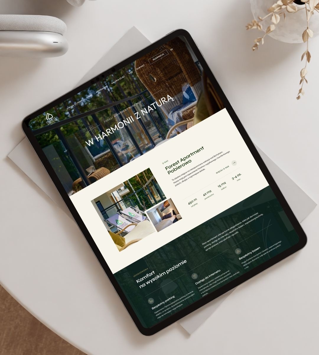 Tablet prezentuje stronę internetową Forest Apartment Pobierowo z hasłem 'W harmonii z naturą'. Widoczne udogodnienia: parking, internet, basen.