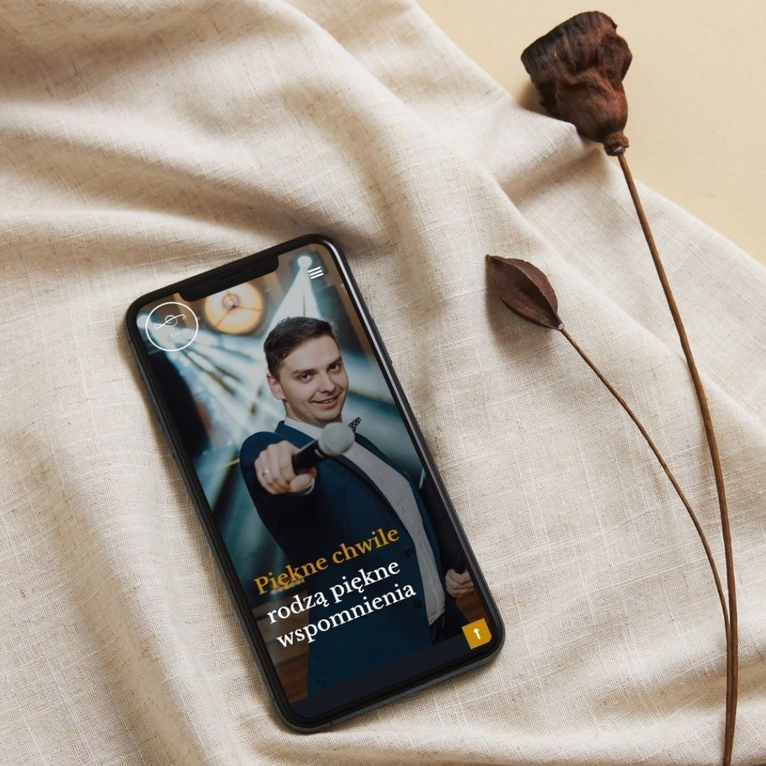 Smartfon wyświetla stronę z uśmiechniętym mężczyzną z mikrofonem, na tle beżowej tkaniny i suszonych kwiatów. Minimalistyczny design strony z hasłem o pięknych chwilach.