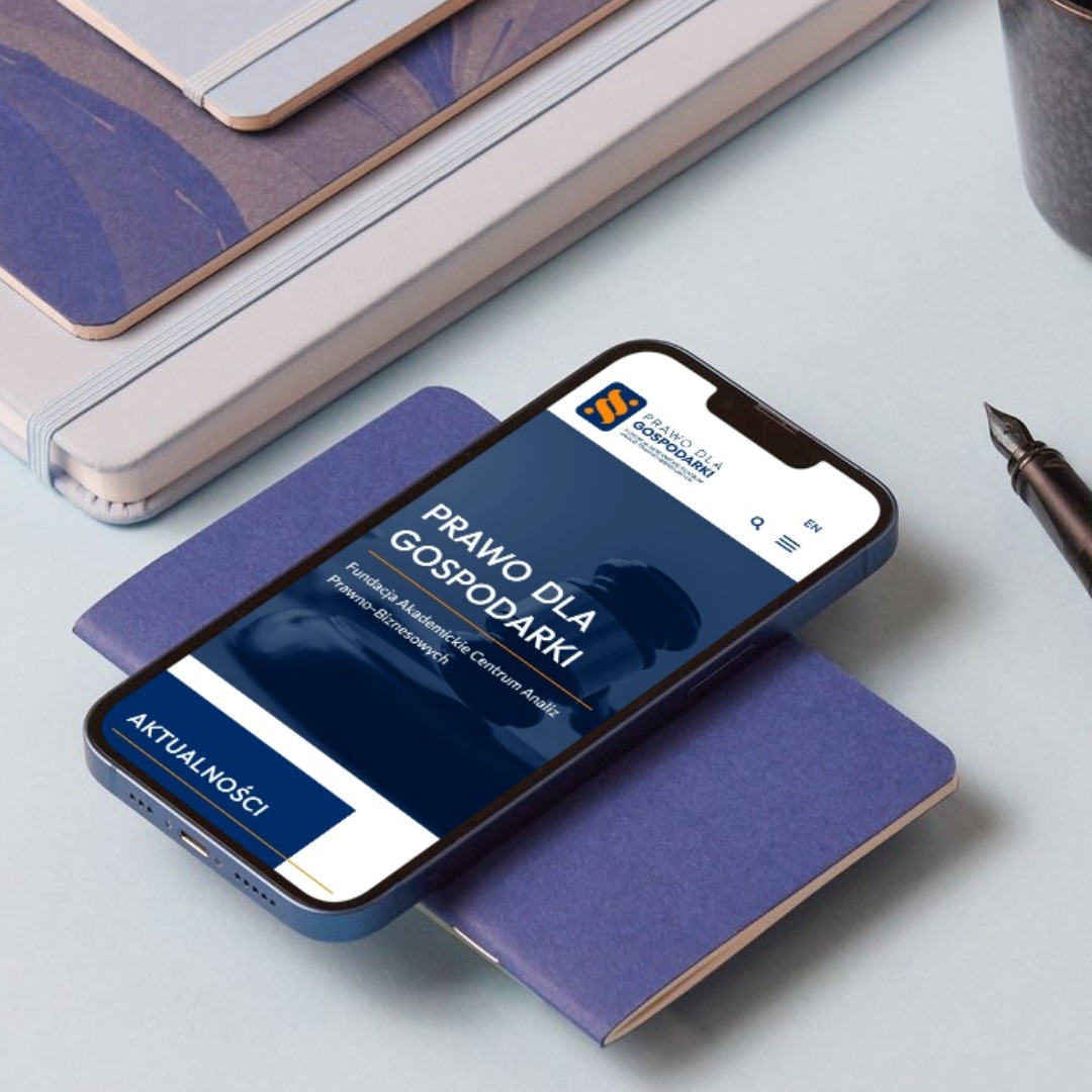 Smartfon prezentuje stronę internetową 'Prawo dla Gospodarki' na tle notatników i pióra. Minimalistyczny design, biznesowy charakter, nowoczesny interfejs.
