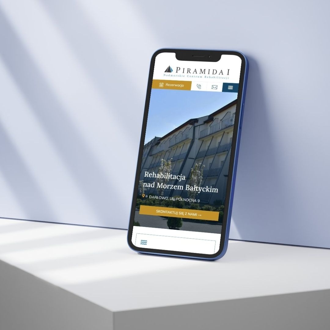Smartfon z wyświetloną mobilną wersją strony internetowej ośrodka rehabilitacyjnego 'Piramida I' na tle białego blatu i błękitnej ściany.