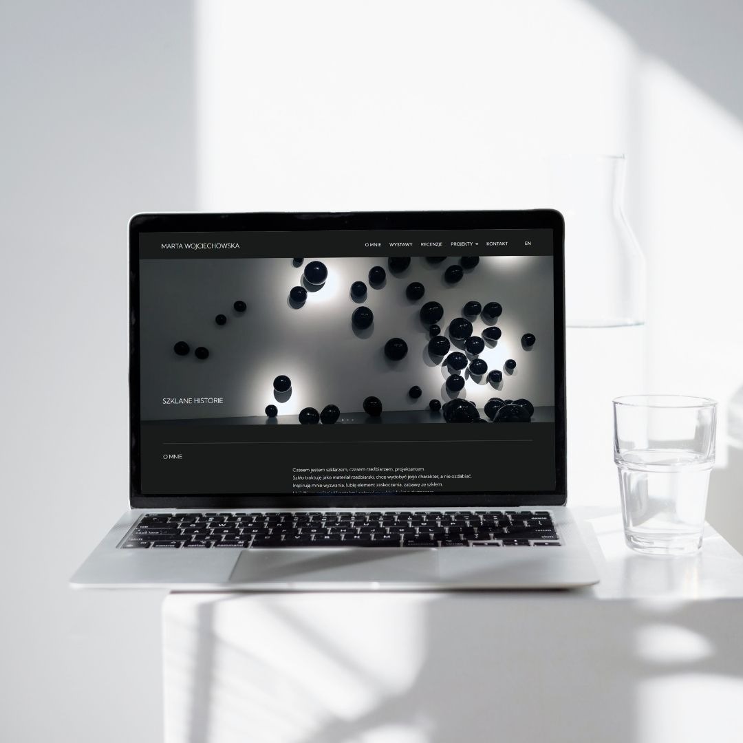Laptop z otwartą stroną internetową artystki Marty Wojciechowskiej, prezentującą szklane rzeźby. Minimalistyczny design, szklanka wody obok.