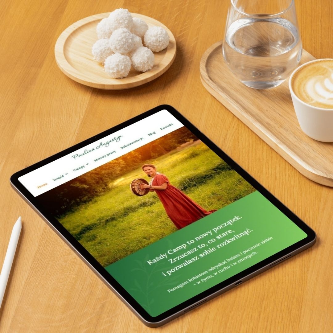 Tablet prezentuje stronę internetową Pauliny Augustyn z kobietą na łące. Obok kawa, woda i kokosowe kulki na drewnianym stole. Minimalistyczny design, ciepłe światło.
