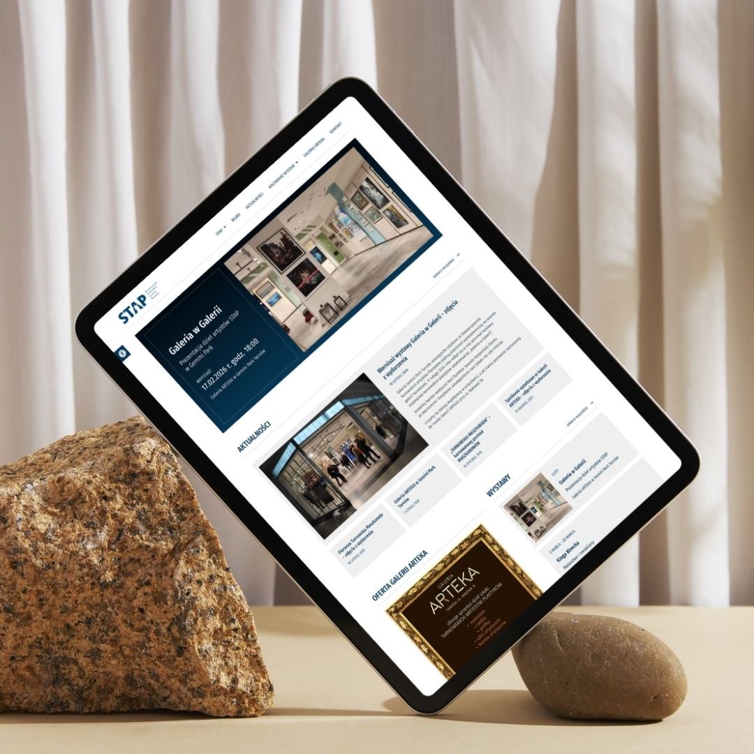 Tablet prezentuje stronę internetową galerii sztuki STAP, z aktualnościami i ofertą Galerii Arteka. Ustawiony na kamieniach, na tle kremowej tkaniny.