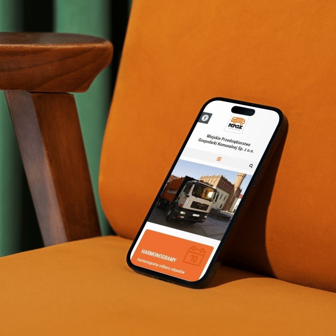 Smartfon prezentuje stronę internetową MPGKi harmonogram odbioru odpadów na pomarańczowym fotelu. Widok z boku, detale interfejsu i kolorystyka.