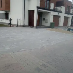 Szara kostka brukowa ułożona przed szeregiem domów jednorodzinnych, widoczny wózek z piaskiem i zagęszczarka wibracyjna, etap prac.