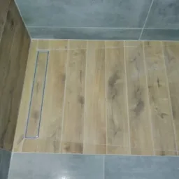 Brodzik wyłożony płytkami imitującymi drewno z odpływem liniowym, obrzeża wykończone szarymi płytkami.