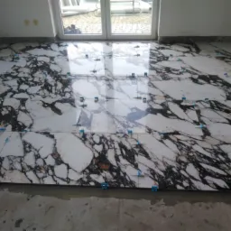 Układanie płytek imitujących marmur na podłodze, z widocznymi niebieskimi krzyżykami dystansowymi, w trakcie prac wykończeniowych, z oknem balkonowym w tle.