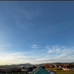 Marek - Panorama z widokiem na górzysty krajobraz i zabudowę mieszkalną. Na pierwszym planie fragment dachu z zieloną membraną i panelami słonecznymi. Błękitne niebo z chmurami.