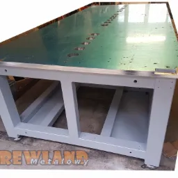 Stół roboczy z metalowym blatem, z szeregiem okrągłych otworów, umieszczony na szarej, metalowej konstrukcji z regulowanymi nóżkami. Widoczny logotyp firmy w dolnej części zdjęcia.