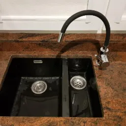 Czarny, dwukomorowy zlewozmywak z baterią z elastyczną wylewką na blacie z czerwonego granitu pod oknem.