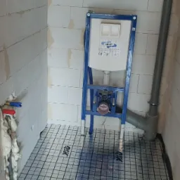 Stelaż podtynkowy WC zamontowany w surowym wnętrzu łazienki z bloków, widoczne rury doprowadzające wodę oraz odpływ kanalizacyjny, na podłodze mata grzewcza.