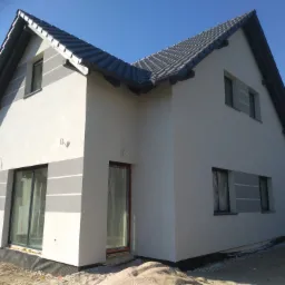 Nowoczesny dom jednorodzinny z jasną elewacją ozdobioną szarymi pasami, ciemnoszarym dachem z dachówki i ciemnymi ramami okiennymi, widoczny z zewnątrz w słoneczny dzień.