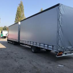 Punkt - Produkcja i Naprawa Plandek - Długi zestaw ciężarowy z szarą plandeką, widoczny pracownik w czerwonym kombinezonie na tle betonowego placu i drzew.