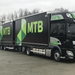 Punkt - Produkcja i Naprawa Plandek - Czarny ciągnik siodłowy DAF XF z naczepą, z dużym logo MTB na burcie, zaparkowany na brukowanym placu, z widocznymi postaciami w oddali.